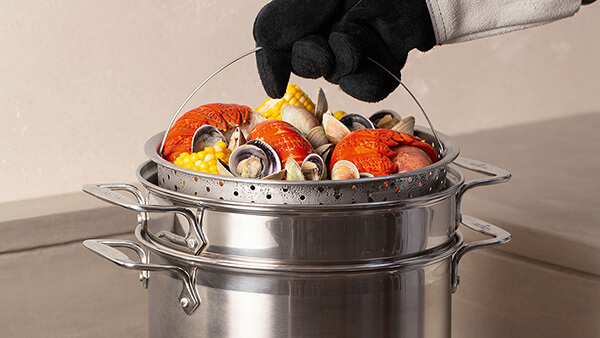 Hand wearing heat resistant glove, holding a steaming basket with boiled seafood